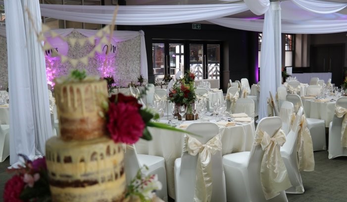 County Long Room dressed for a wedding at Chester Racecourse. Wedding cake is rustic with floral details and gold bows on chair decoration.